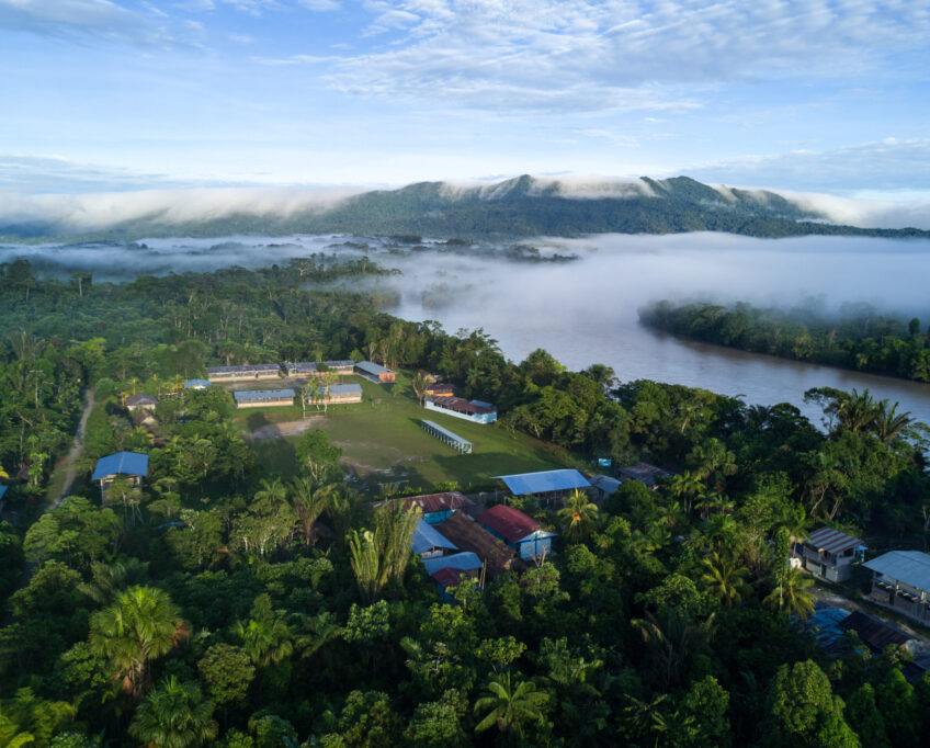 Aerial image of a village near a river with forested mountains in the background.